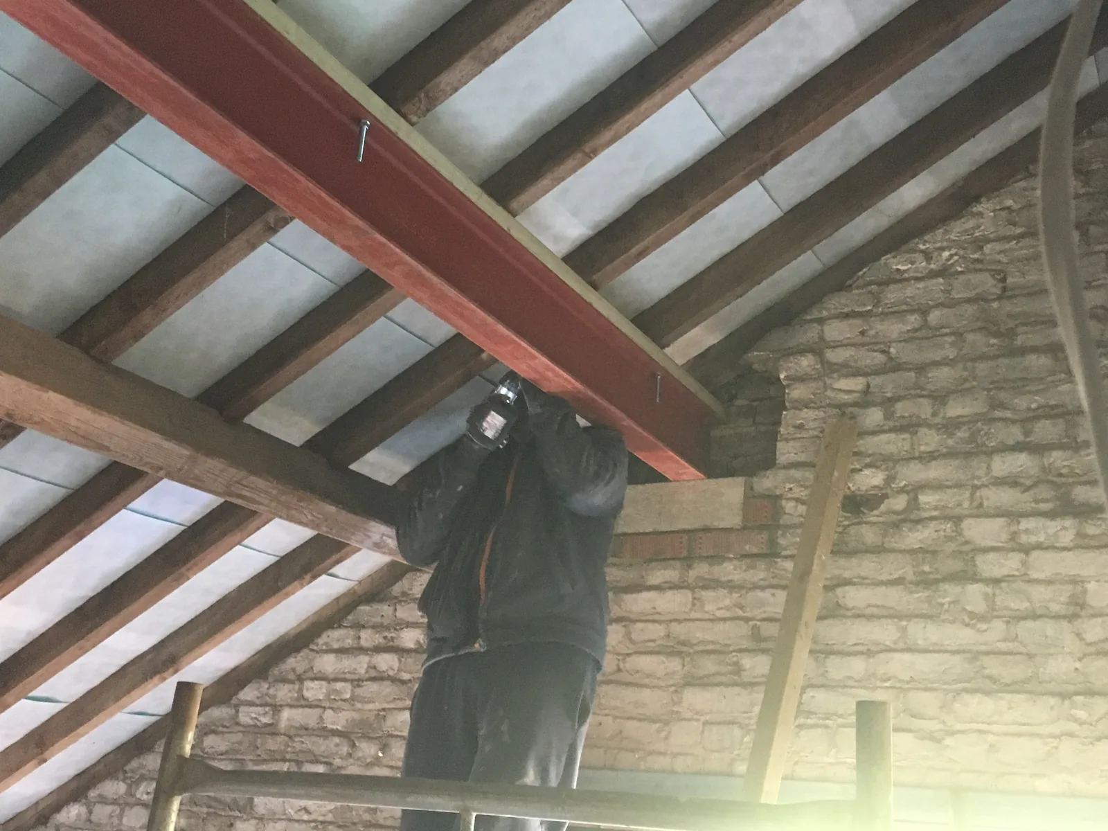 A red-oxide structural steel beam being bolted up under the original rafters, against the bare brick and stone of an internal gable wall.