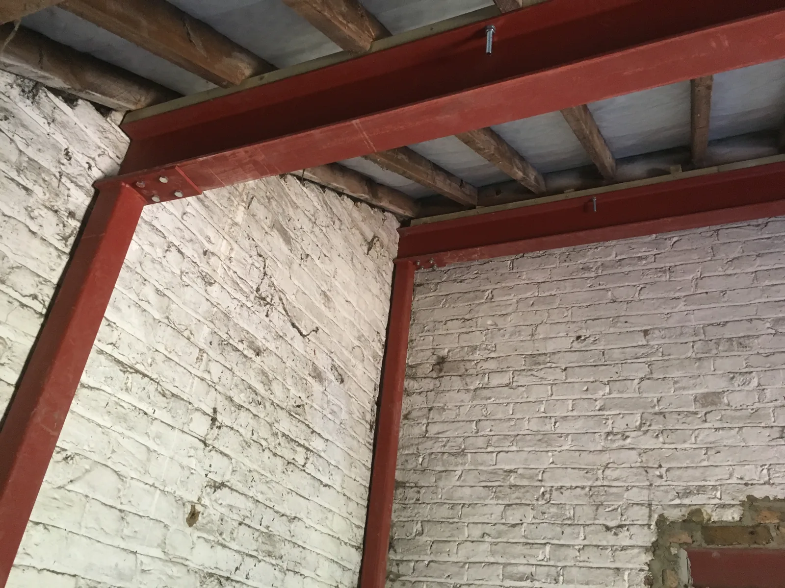 Two red-oxide steel beams and a supporting post bolted together into a frame, seated against whitewashed brick walls with old timber joists overhead.