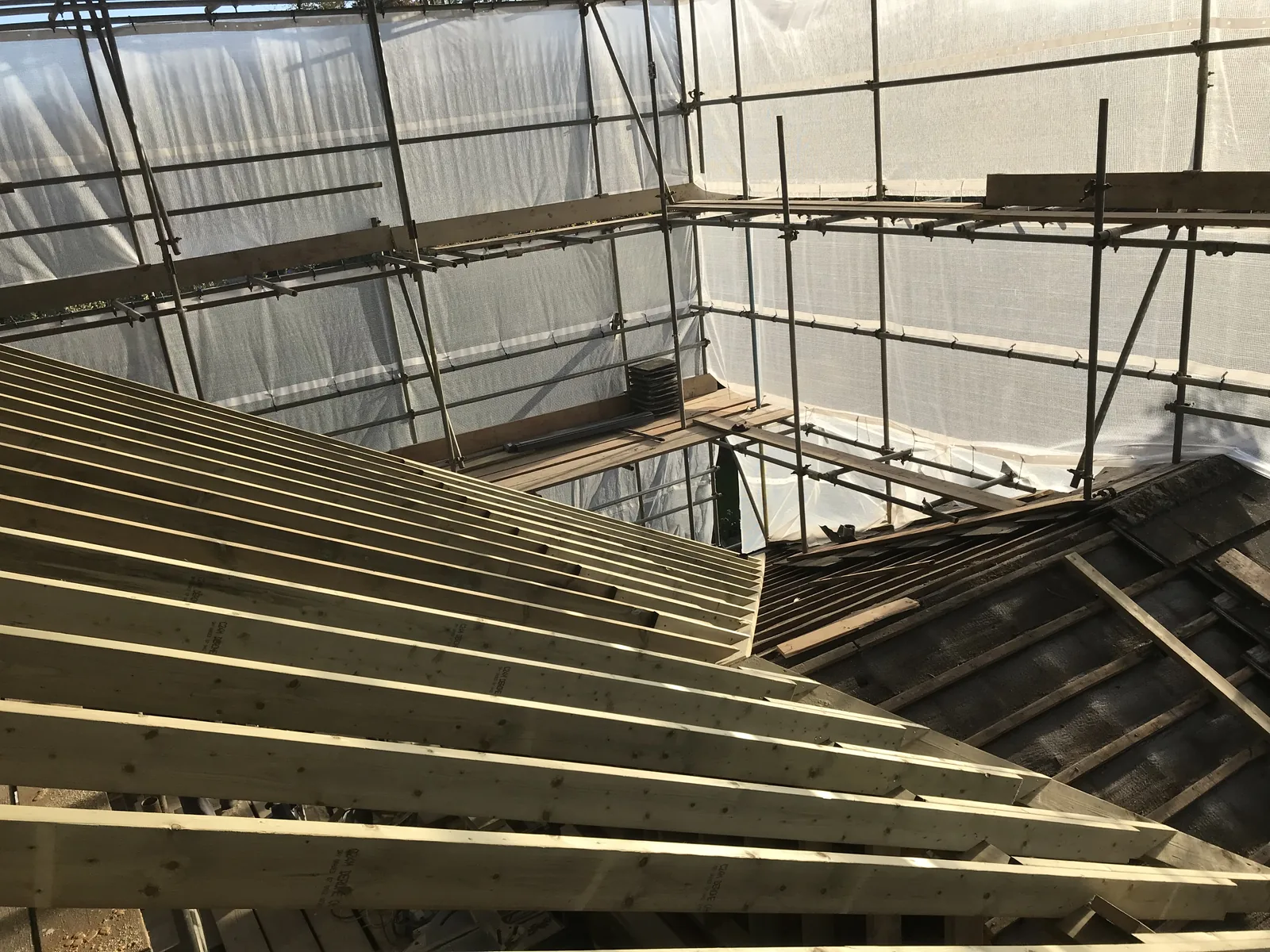 New pale timber rafters rising beside a stripped, felted section of old roof, the whole house wrapped in scaffold and protective sheeting.