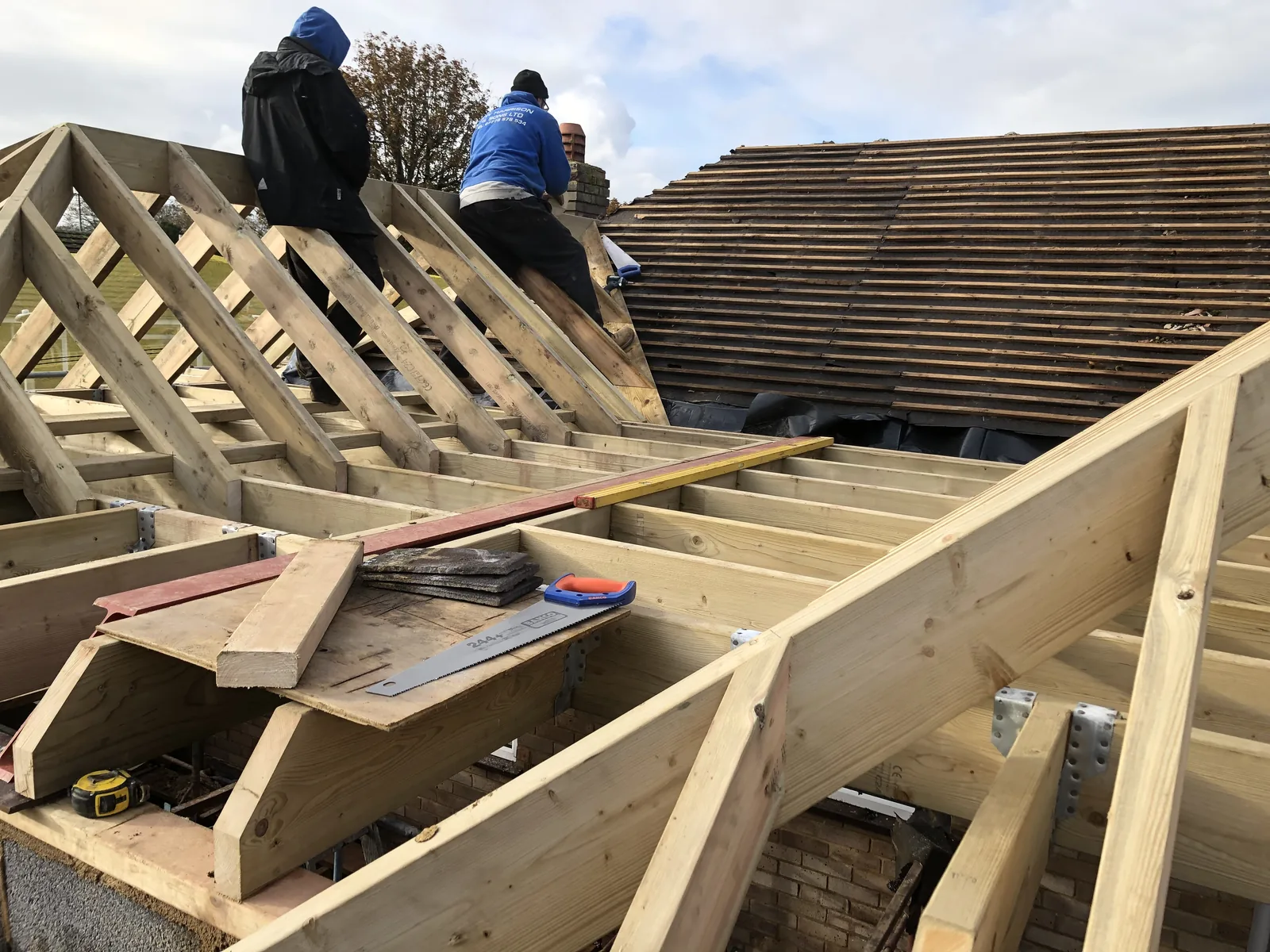 Two builders in branded workwear cutting and fixing timber on the new roof frame, with the stripped batten roof of the existing house alongside them.