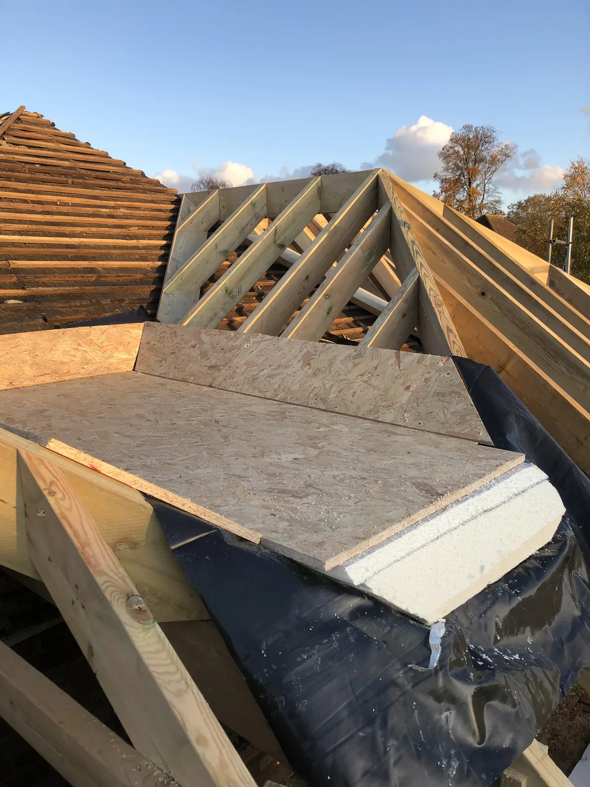 Board decking and rigid insulation laid over a flat section of the new roof on a black membrane, with a new hipped rafter frame behind and old stripped roof beyond.