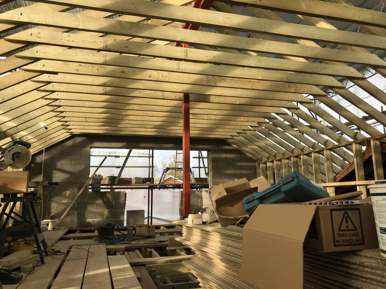 Inside the new attic room, looking along the rafters to a blockwork gable with a large window opening framed into it and a red steel post rising to the ridge.