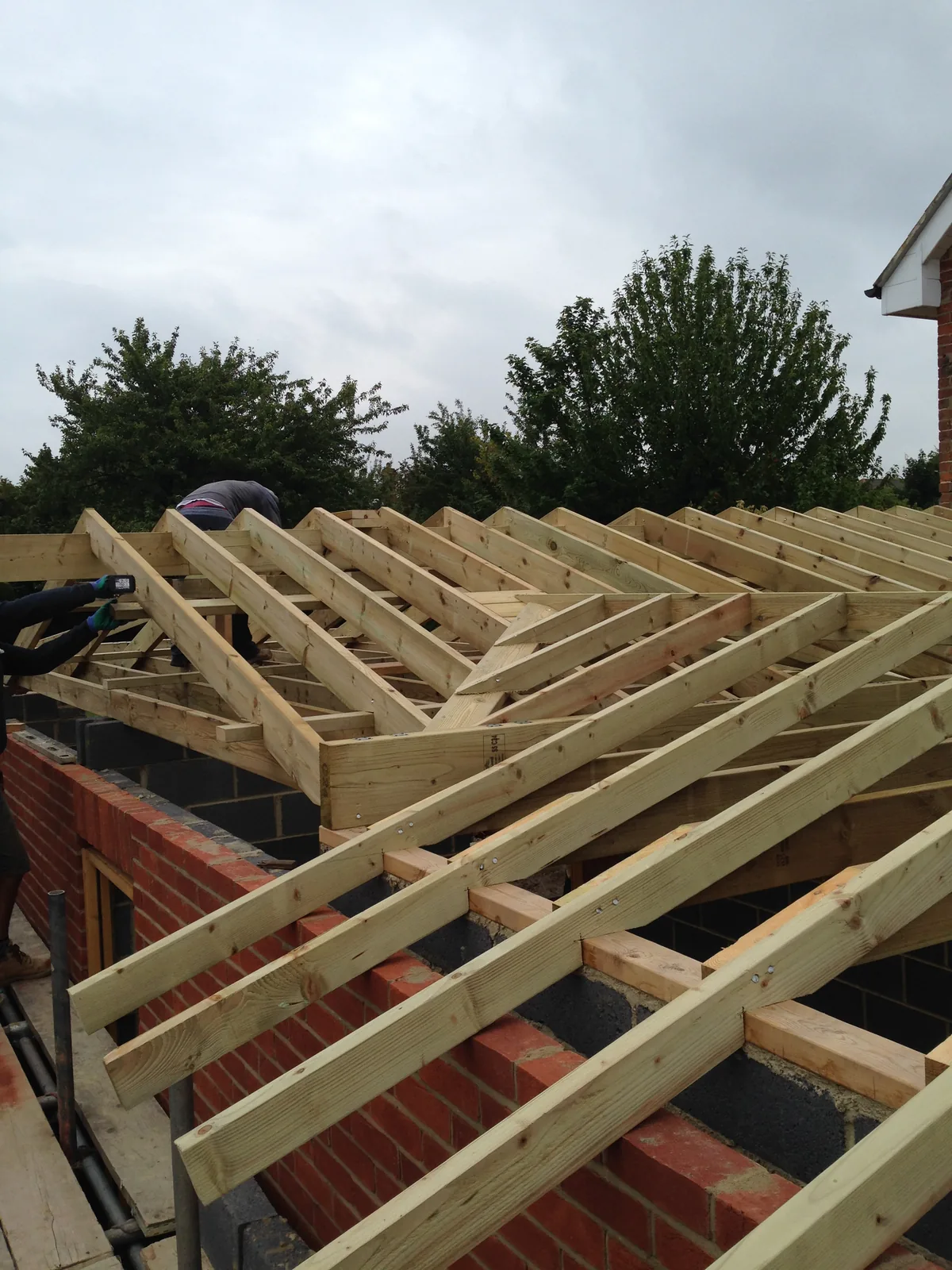 A cut hip roof being assembled on site over new cavity walls, the brick outer leaf and block inner leaf visible below rafters seated on the wall plate.