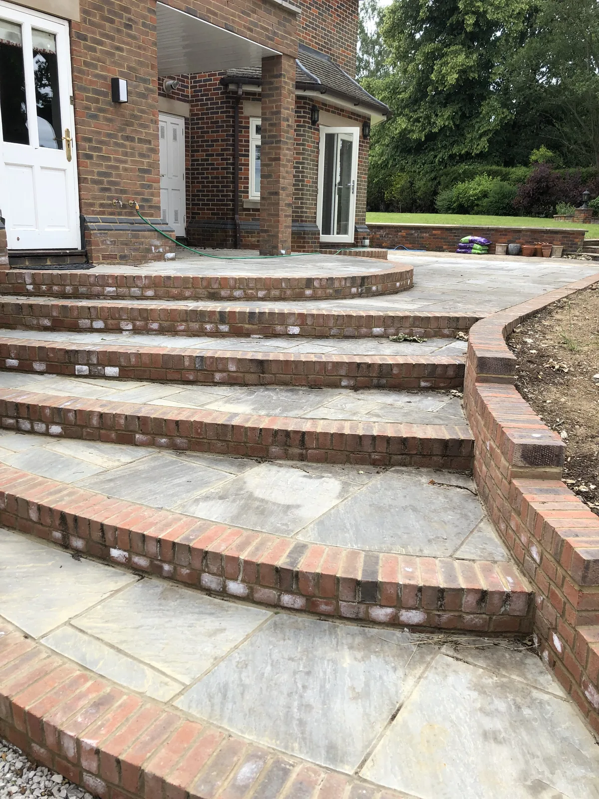 Natural stone terraces stepping down from a brick house on a sweeping curve, each level trimmed with a brick-on-edge riser and a curved brick retaining wall to one side.